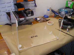 Clear acrylic box or container with metal components inside, sitting on wooden surface in workshop or lab setting