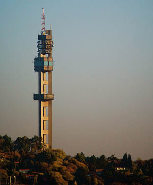 Tall concrete telecommunications tower with antennas on hilltop, surrounded by trees under evening sky