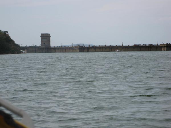 View from boat of large concrete dam wall with control tower across choppy water under overcast sky