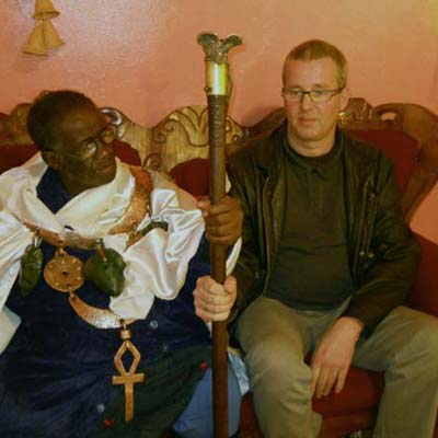 Two men sitting on couch indoors, one Black man in white shirt with medallion, one white man in dark jacket holding decorative staff