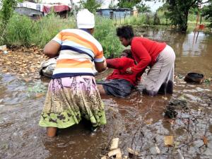 Two people kneeling in muddy floodwater, one wearing striped shirt and patterned skirt, another in red shirt, houses visible