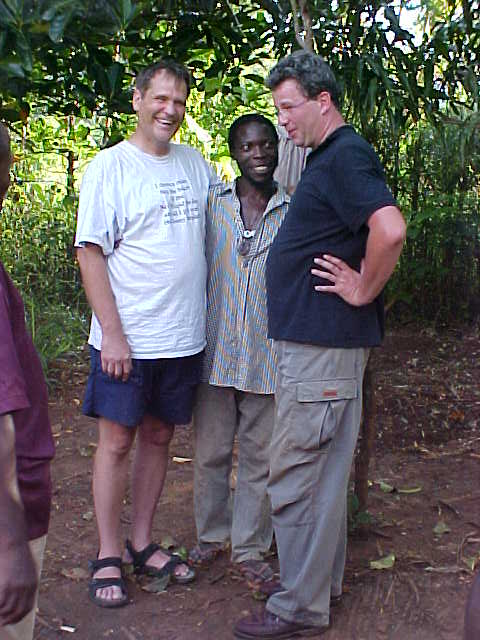 Three men posing together outdoors on dirt ground with lush green foliage and trees in background
