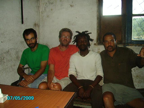 Four men sitting together on benches indoors, wearing casual colored shirts, with white walls and windows visible behind them