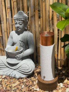 Gray Buddha statue holding yellow flower petal sits beside white/copper cylindrical device on rocky ground with bamboo fence