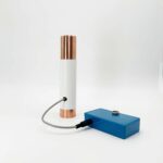White cylindrical tube with copper-colored metal ends connected by gray cable to blue rectangular control box on white background
