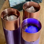 Three copper-colored metal cups with purple leather sleeves, containing dark liquid and ice cubes, one glowing blue from below