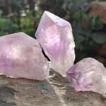 Three translucent purple amethyst crystal chunks on weathered stone surface, outdoor natural lighting with blurred greenery