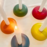 Five colorful circular orgonite candle holders with white candles - green, red, orange, yellow, and blue/purple on white surface