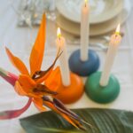 Orange bird of paradise flower next to three round orgonite candle holders in orange, blue, and green with lit white candles