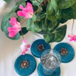 Quatre dessous-de-verre ronds bleus pailletés avec centres métalliques spiralés, un tenant un verre avec glace, entourés de fleurs roses
