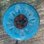 Round blue orgonite disc with metallic spiral center and crystal points, viewed from above on textured surface