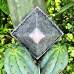Pirámide orgonite gris oscuro con centro de cuarzo rosa, colocada sobre cactus verdes rodeados de pequeñas plantas verdes y amarillas