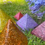 Colorful orgonite pyramids on grass: large amber pyramid in foreground, purple and pink squares behind, with purple flowers