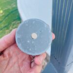 Hand holding circular blue-gray orgonite disc with central metal piece, showing textured surface with embedded materials