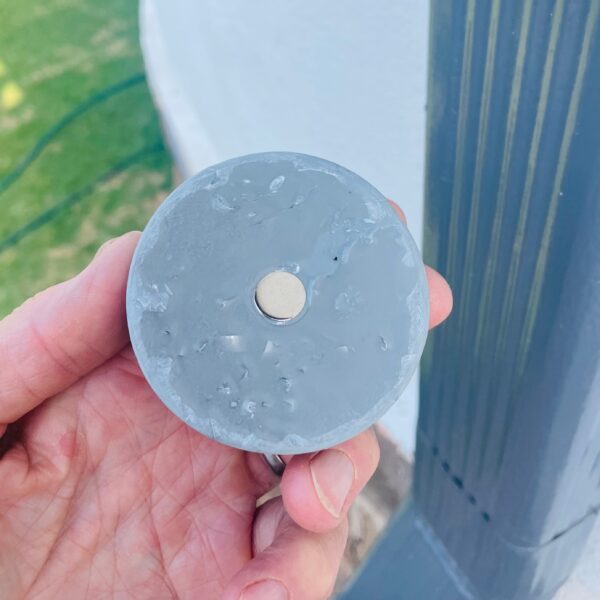 Hand holding circular blue-gray orgonite disc with central metal piece, showing textured surface with embedded materials