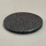 Black circular disc with rough, textured surface containing white speckles, photographed from above on white speckled background