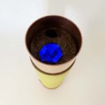 Cylindrical container with yellow bottom, brown middle band, and brown interior containing dark material and bright blue object