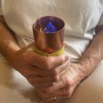 Hands holding copper cylinder with yellow band and blue crystal inside, worn on yoga mat