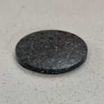 Disco circular negro con superficie metálica moteada sobre encimera blanca jaspeada, visto desde arriba en ángulo