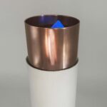Kupferfarbener zylindrischer Metallbehälter mit dunklem Innenraum, blaues dreieckiges Objekt innen sichtbar, auf reflektierender Oberfläche