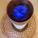 Zylindrisches kupferumrandetes Rohr mit weißer Basis, dunkler Körnerschicht und blauem Kristallwürfel oben, von oben auf Holz