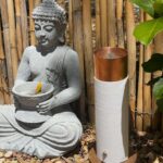 Weißer zylindrischer orgonite Stab mit Kupfer-Oberteil auf Holzbasis, neben grauer Buddha-Statue mit gelber Blume vor Bambuszaun