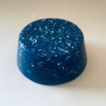 Cilindro orgonite azul con virutas metálicas incrustadas, fotografiado desde arriba sobre superficie blanca