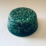 Disco orgonite verde redondo con motas metálicas incrustadas, fotografiado desde arriba sobre fondo beige