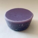 Pieza de orgonite violeta en forma de cúpula con superficie inferior texturizada, fotografiada en ángulo sobre fondo beige