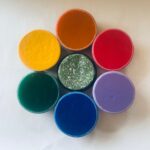 Seis piezas circulares de orgonite dispuestas en patrón floral sobre superficie blanca. Colores: amarillo, naranja, rojo, morado, azul, verde con copos blancos.