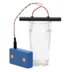 Dispositivo rectangular azul con círculos de cobre conectado por cables rojo-negro a electrodo de barra negra en vaso de agua