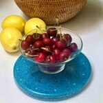 Blue circular charging plate with glass bowl of red cherries on top, three yellow lemons beside it, woven basket in background