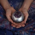 Mains tenant un objet sphérique métallique avec des cristaux violets au sommet, sur un tissu à motifs paisley bleus
