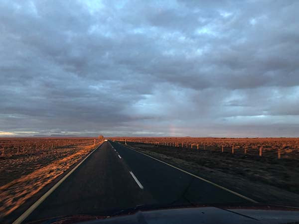 Straight asphalt road stretching to horizon through arid reddish landscape under dramatic cloudy sky at sunset/sunrise