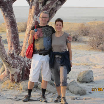 Two people posing together under a large tree in arid landscape with scattered rocks and sparse vegetation